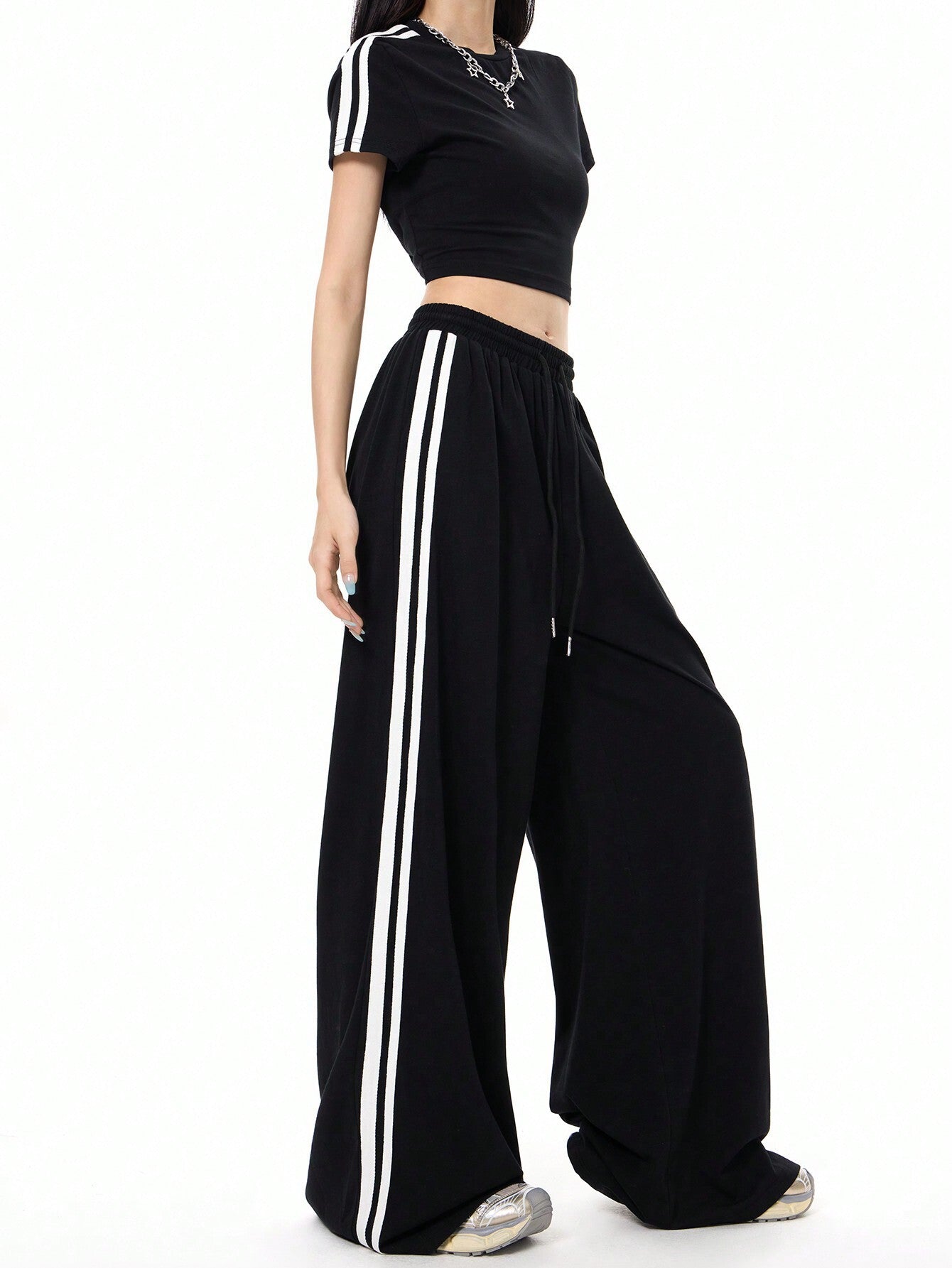 A woman is standing in profile view showcasing a black athletic two-piece outfit with white side stripes and ultra-wide palazzo-style pants