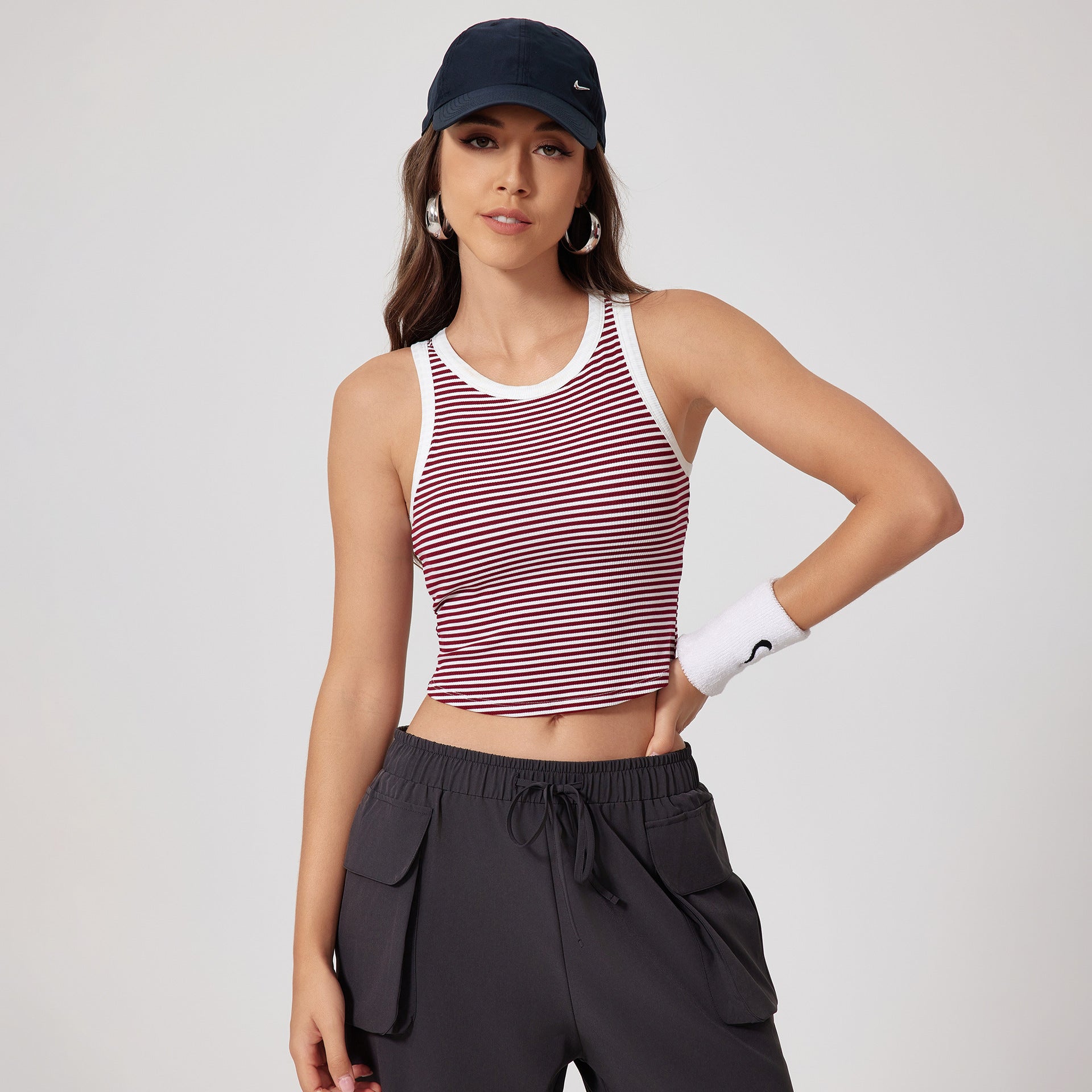 A woman wearing wine red striped crop top with white trim, navy cap, white wristband and gray cargo pants against clean white background