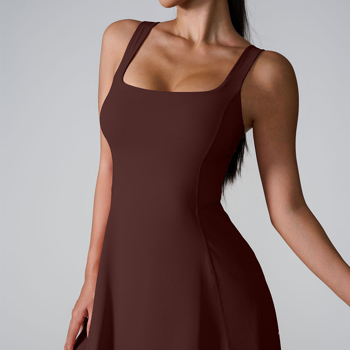 A model in chocolate brown square neck dress with structured fit and wide shoulder straps against light gray setting