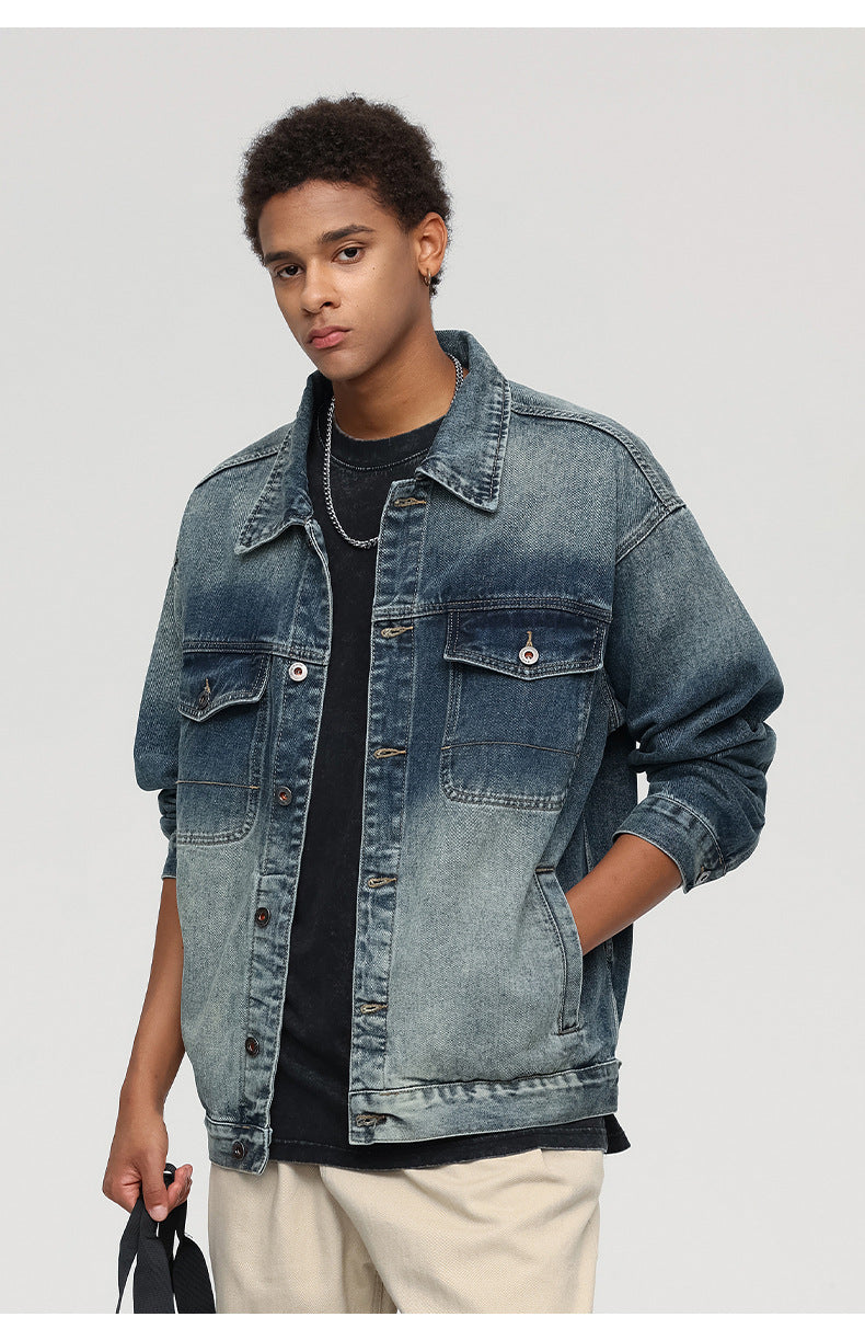 A man is standing with one hand holding fabric against a gray backdrop. Wearing gradient blue denim jacket with visible chest pockets and beige pants