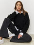 Woman seated wearing black sweater layered over white collared shirt with black pants and white sneakers