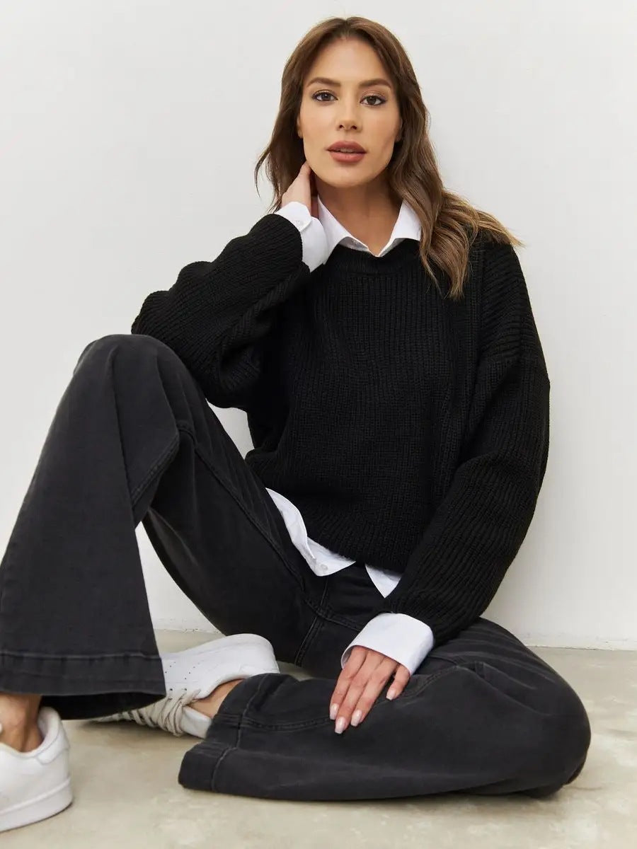 Woman seated wearing black sweater layered over white collared shirt with black pants and white sneakers