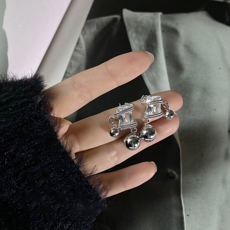 A hand holding silver dangle earrings with large clear gemstones and small metallic ball charms, against a dark background.