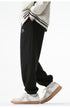 A model is wearing black sweatpants with an elastic waistband, loose fit, and subtle logo detailing, standing with one hand in the pocket