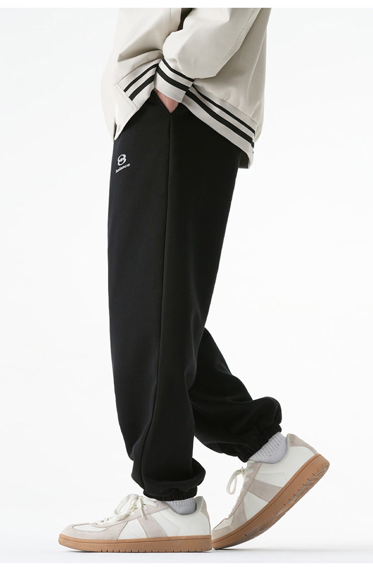 A model is wearing black sweatpants with an elastic waistband, loose fit, and subtle logo detailing, standing with one hand in the pocket
