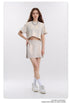 Model wearing cropped beige t-shirt and matching mini skirt with silver chain necklace, white socks and sneakers against white background