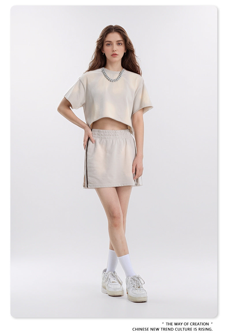 Model wearing cropped beige t-shirt and matching mini skirt with silver chain necklace, white socks and sneakers against white background