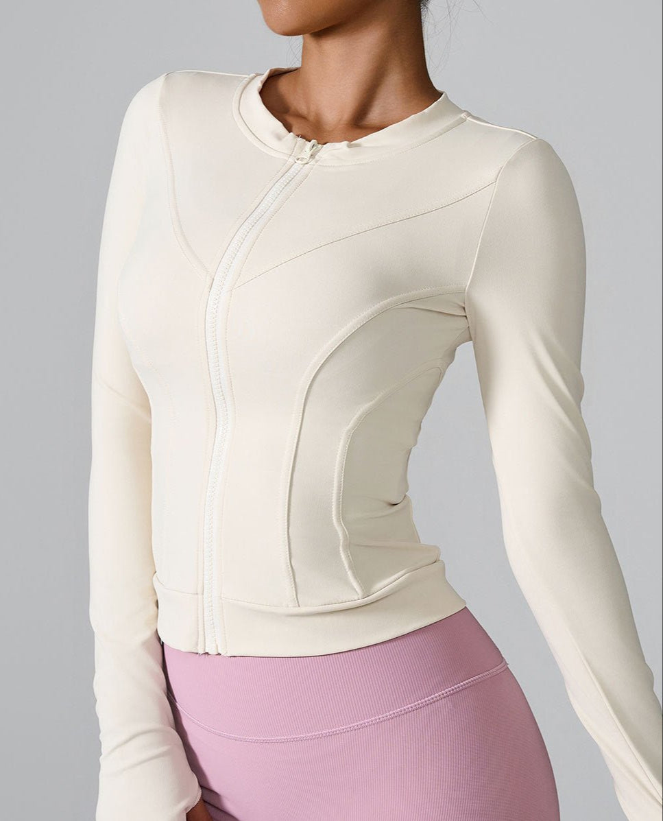 A model is wearing a cream fitted athletic jacket with front zip closure and contoured seams against gray background. Paired with pink leggings