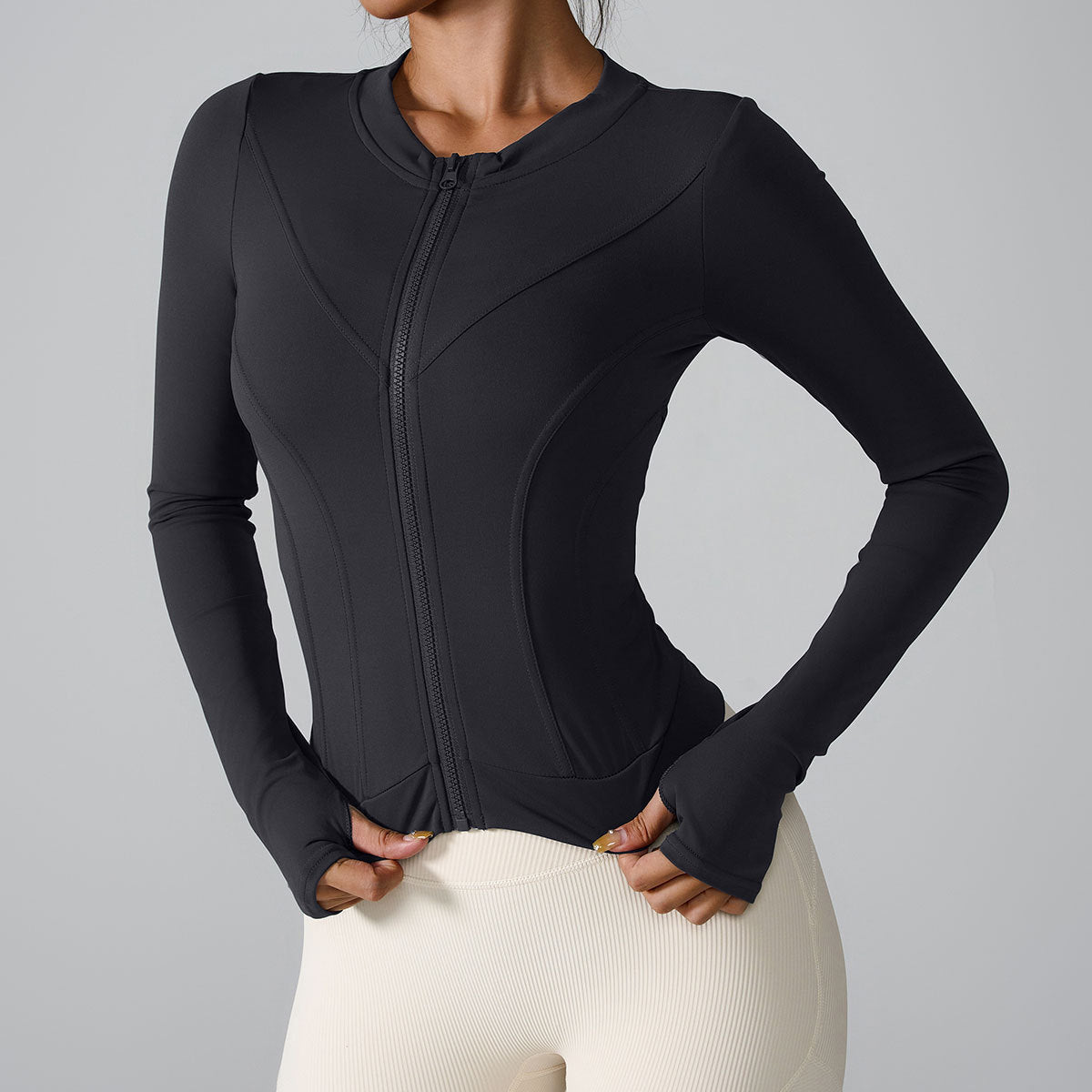 A model is wearing a black performance jacket with technical seaming and front zip closure. Hands positioned at waistband of cream leggings
