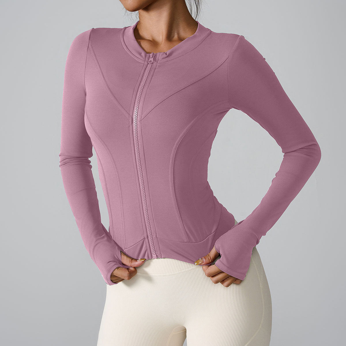 A model is wearing a mauve zip-front athletic jacket with contoured panels paired with cream leggings. Functional design with hand-covering sleeves