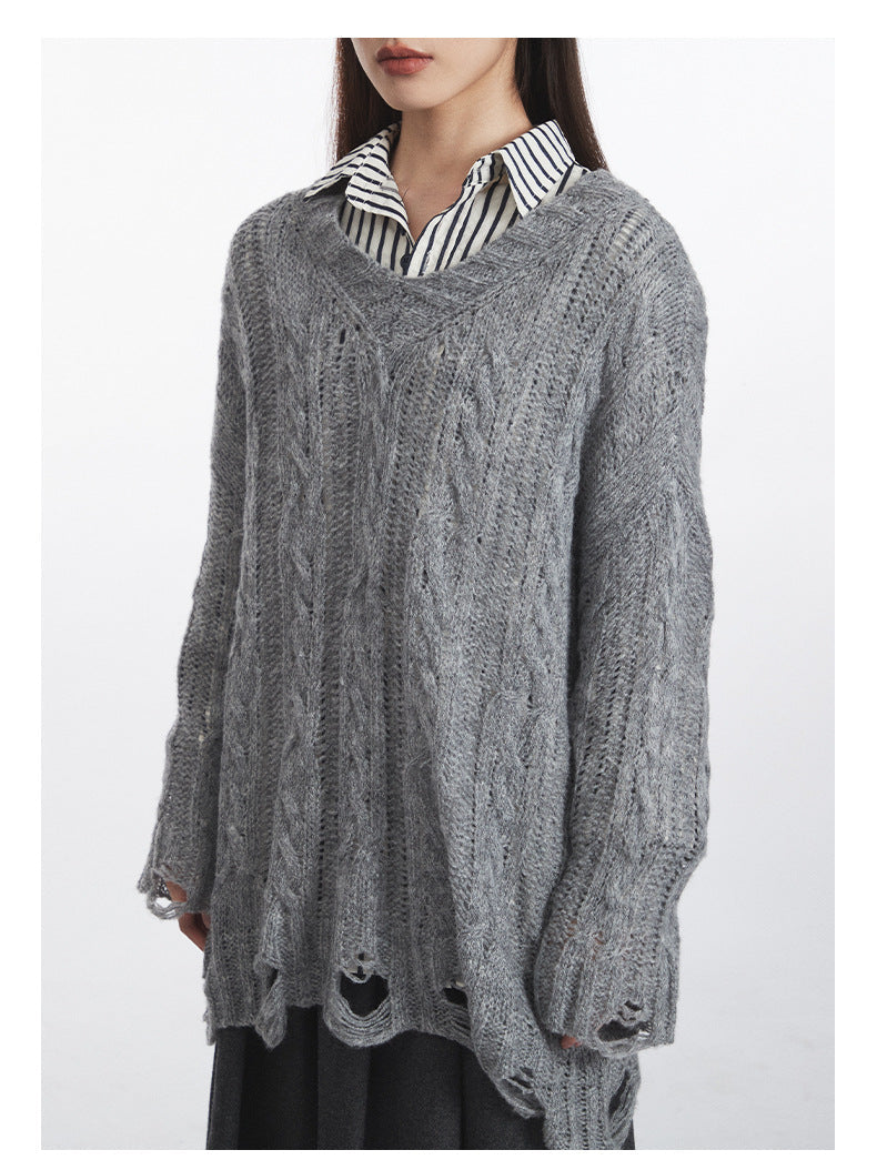 Detailed shot of oversized grey sweater showing V-neck design, cable-knit texture, and distressed bottom edge. Contrasting black and white striped shirt collar visible