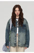 A woman is standing directly facing camera against gray background. Wearing oversized faded denim jacket, white graphic tee and carrying gray shoulder bag