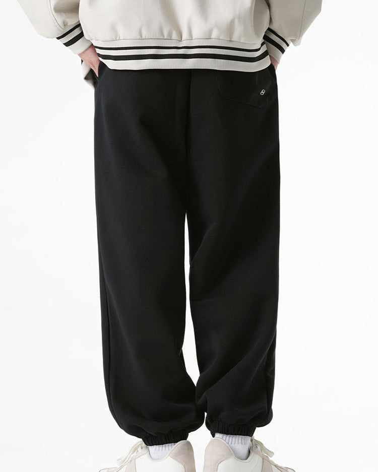 A model is wearing black jogger sweatpants with a single back pocket and elastic cuffs, shown from the rear while paired with a cream varsity jacket and white sneakers