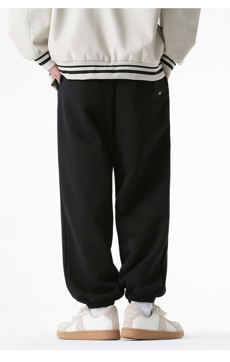 A model is wearing black jogger sweatpants with a single back pocket and elastic cuffs, shown from the rear while paired with a cream varsity jacket and white sneakers