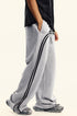 Close-up side view of wide-leg gray sweatpants with black double stripe detail along sides, shown with hand in pocket and black tee