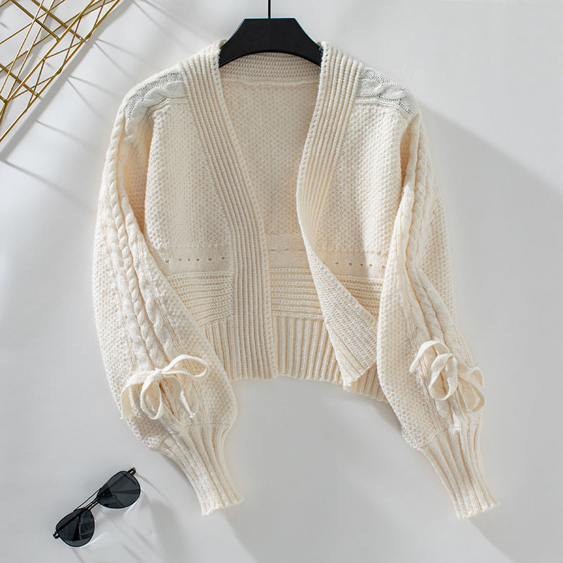 A cream cropped cardigan with cable knit pattern and tie sleeve details displayed on hanger against white background