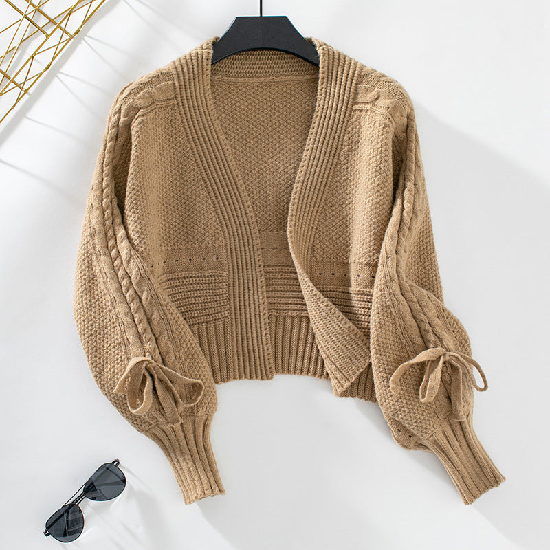 A tan cropped cardigan with ribbed trim and decorative bow sleeve details shown on hanger against minimalist backdrop