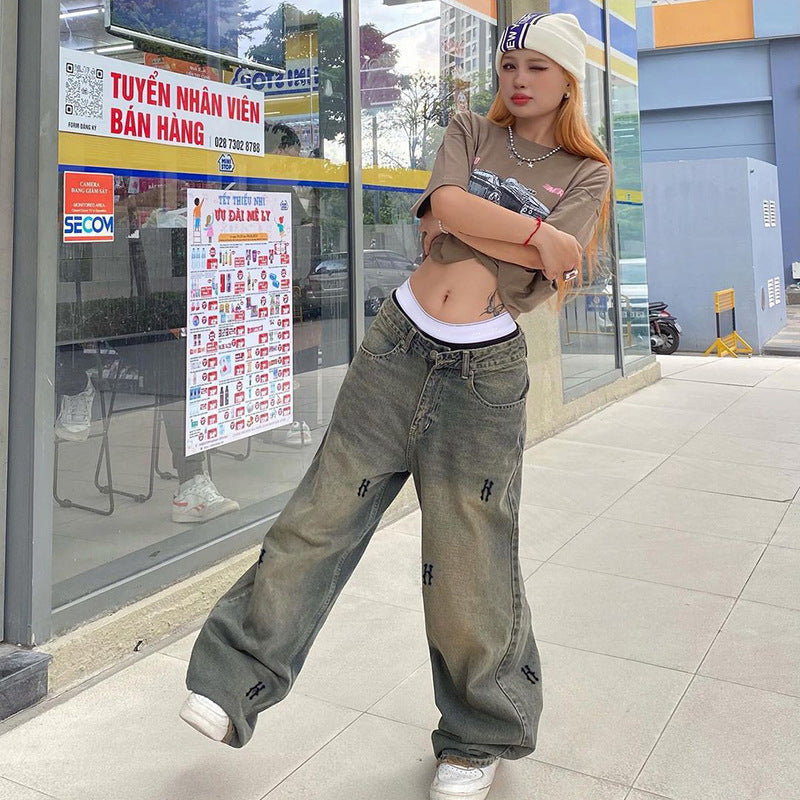 A woman stands with arms crossed outside a store, wearing brown cropped tee, oversized gray jeans with logo details, and white sneakers