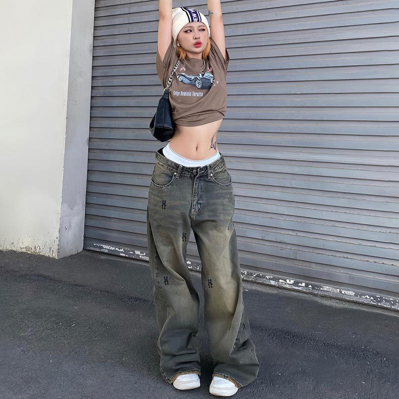 A woman poses with arms raised, wearing cropped brown tee and wide-leg gray jeans with embroidered details, against metal shutters