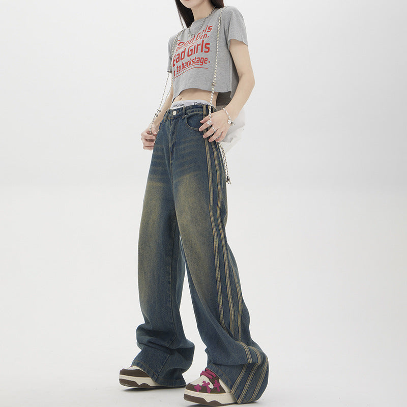 Woman wearing grey 'Bad Girls' crop top, wide-leg jeans with side stripe detail and platform sneakers, carrying chain strap bag against white background