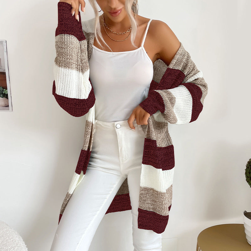 A woman wearing burgundy, beige and white striped long cardigan with white tank top and white pants against white wall
