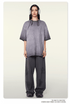 Model wearing oversized gray t-shirt with ombre effect, white headphones, silver chain necklace, and matching wide-leg pants against neutral background