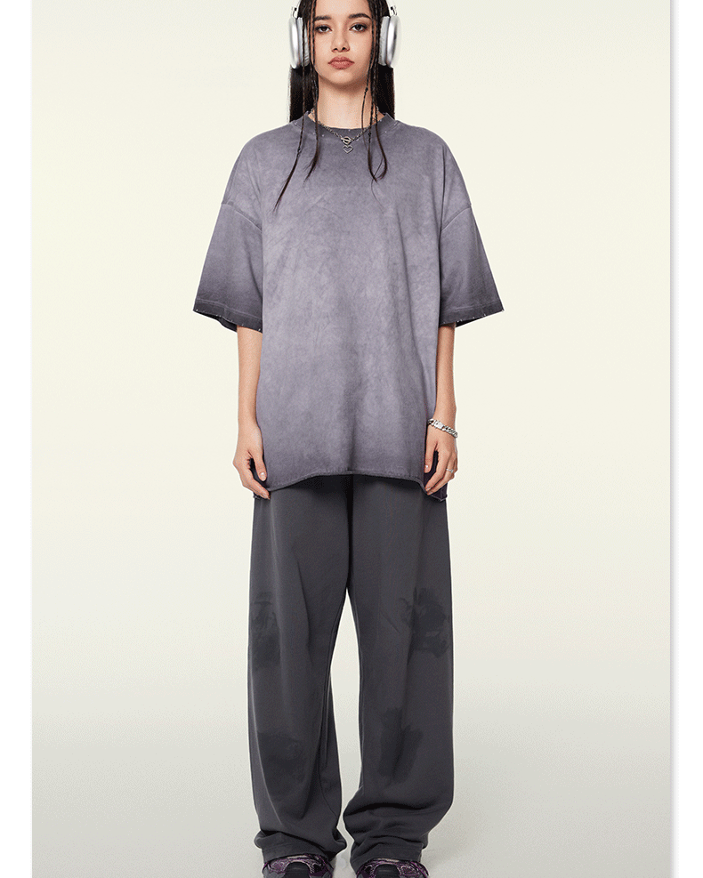 Model wearing oversized gray t-shirt with ombre effect, white headphones, silver chain necklace, and matching wide-leg pants against neutral background