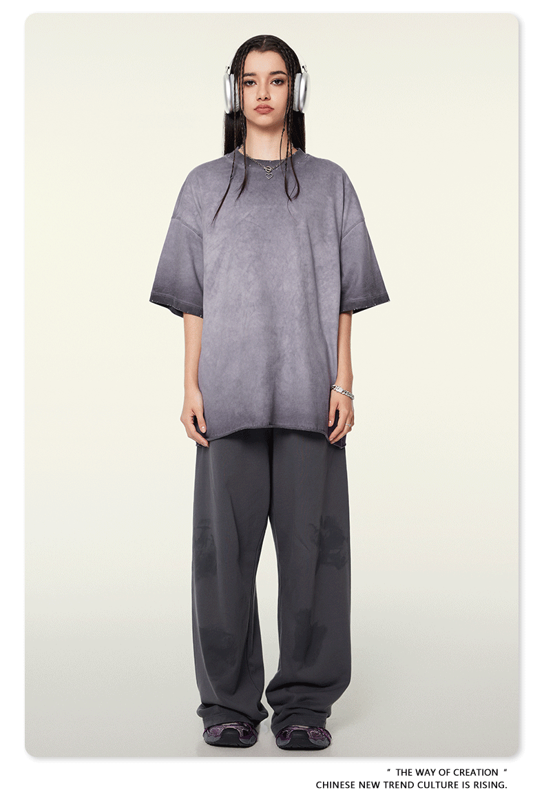 Model wearing oversized gray t-shirt with ombre effect, white headphones, silver chain necklace, and matching wide-leg pants against neutral background