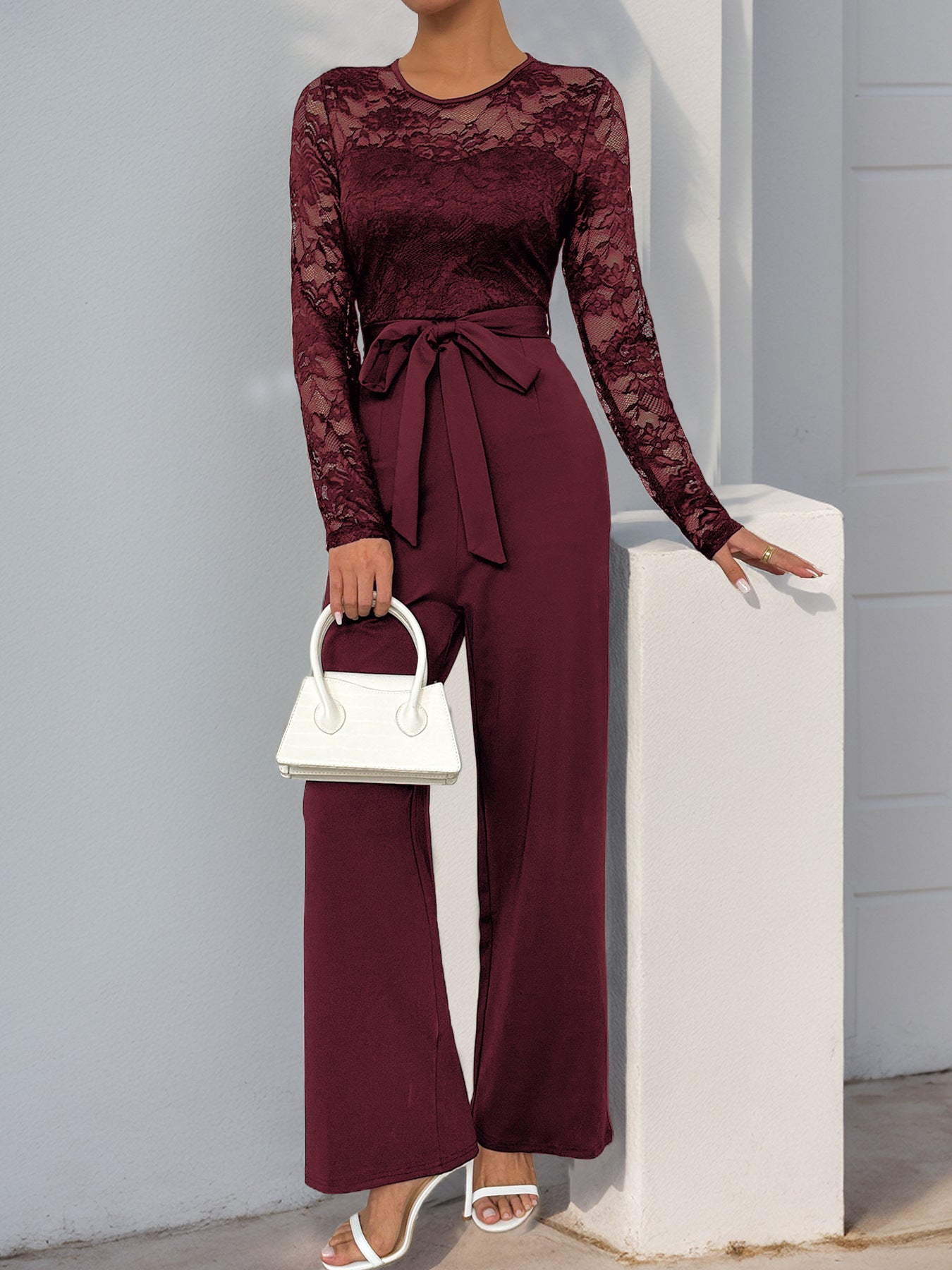 A burgundy version of the lace jumpsuit, styled with white heels and a handbag against a neutral background.