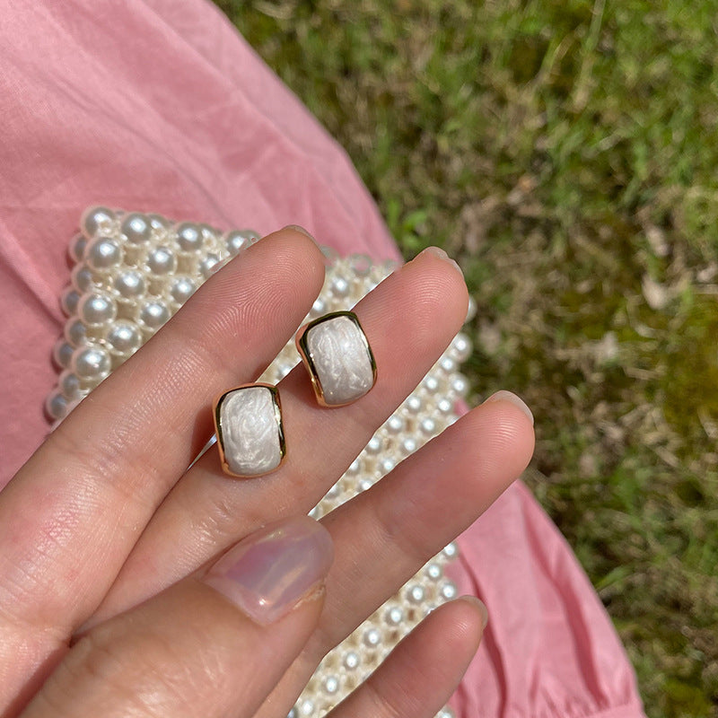 A hand holding a pair of rectangular gold-framed earrings with an irregular white pearl-like texture, set against a pink fabric and pearl handbag.