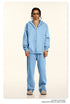 Model wearing oversized light blue zip-up hoodie and matching sweatpants with gold-framed sunglasses and layered silver chain necklaces against cream background