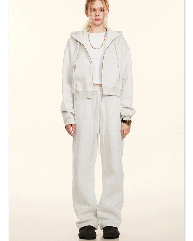 Model standing in white lounge set with cropped zip hoodie and wide-leg sweatpants. Styled with sunglasses and necklace