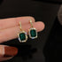 Close-up of emerald and diamond drop earrings held in hand, showcasing rectangular gemstones and sparkling pavé setting against dark background