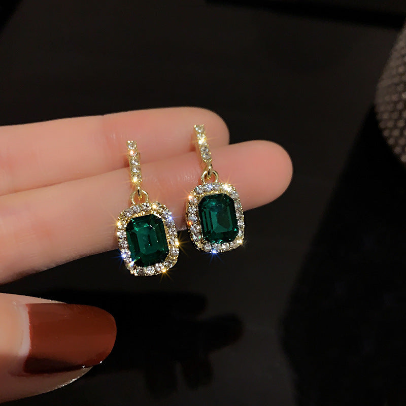 Close-up of emerald and diamond drop earrings held in hand, showcasing rectangular gemstones and sparkling pavé setting against dark background