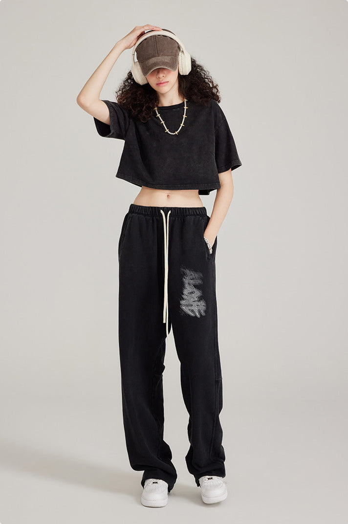 Model in black crop top and black sweatpants with graphic print, styled with white headphones, cap and chunky chain necklace