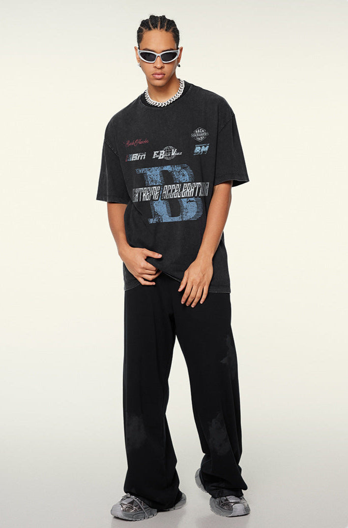 A model posing in black graphic t-shirt with blue 'B' logo design, silver chain necklace, white sunglasses and black wide-leg pants