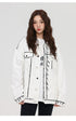 A woman is looking directly at camera. Wearing white SANHOMME outline denim jacket with black contrast stitching over white tee against gray backdrop