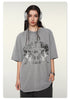 Close-up of model wearing gray oversized t-shirt with reflective "BRAVE ABLE" tiger graphic print, silver headphones and jewelry against white background