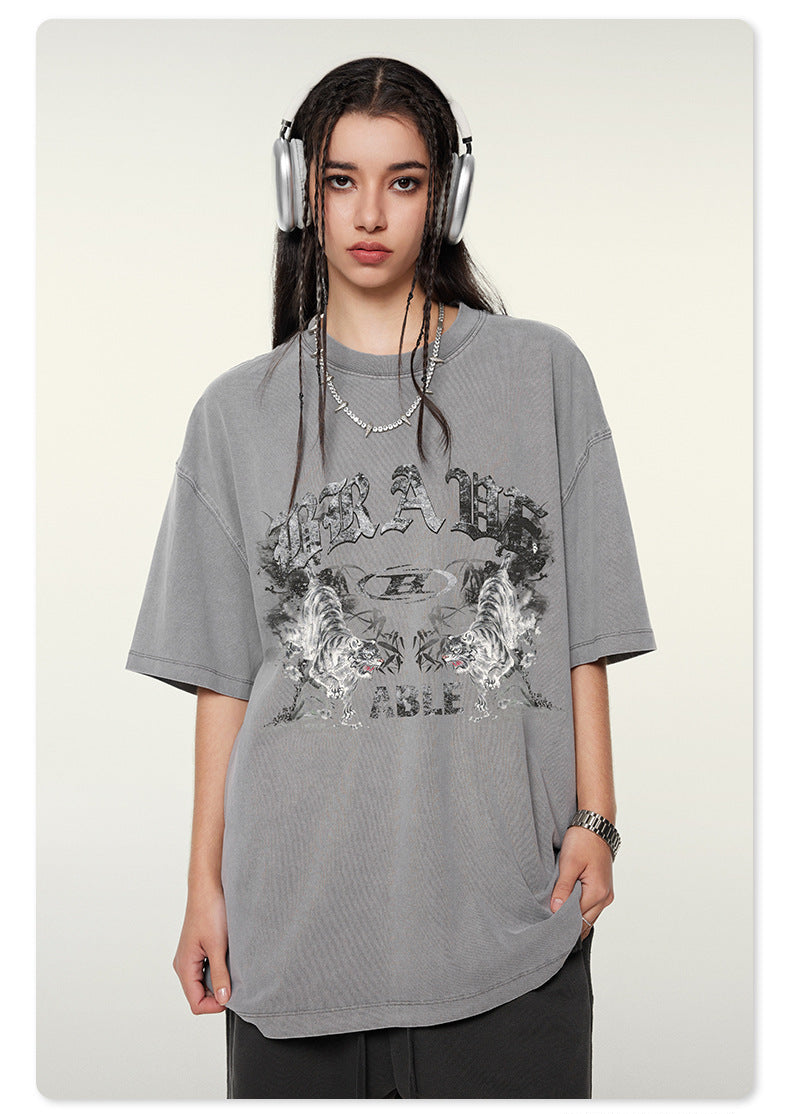 Close-up of model wearing gray oversized t-shirt with reflective "BRAVE ABLE" tiger graphic print, silver headphones and jewelry against white background