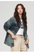 A woman is looking at camera while posing at angle against gray background. Wearing faded denim jacket, white graphic crop top and beige pants