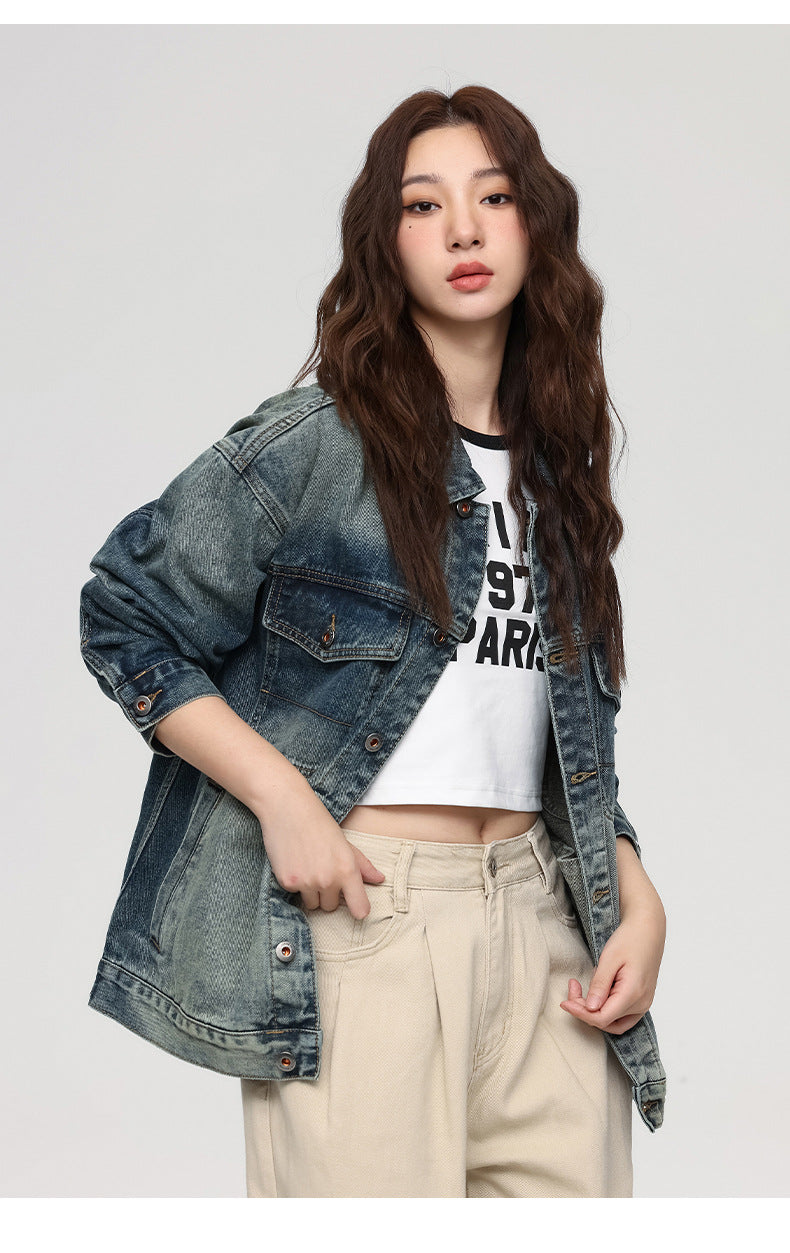 A woman is looking at camera while posing at angle against gray background. Wearing faded denim jacket, white graphic crop top and beige pants