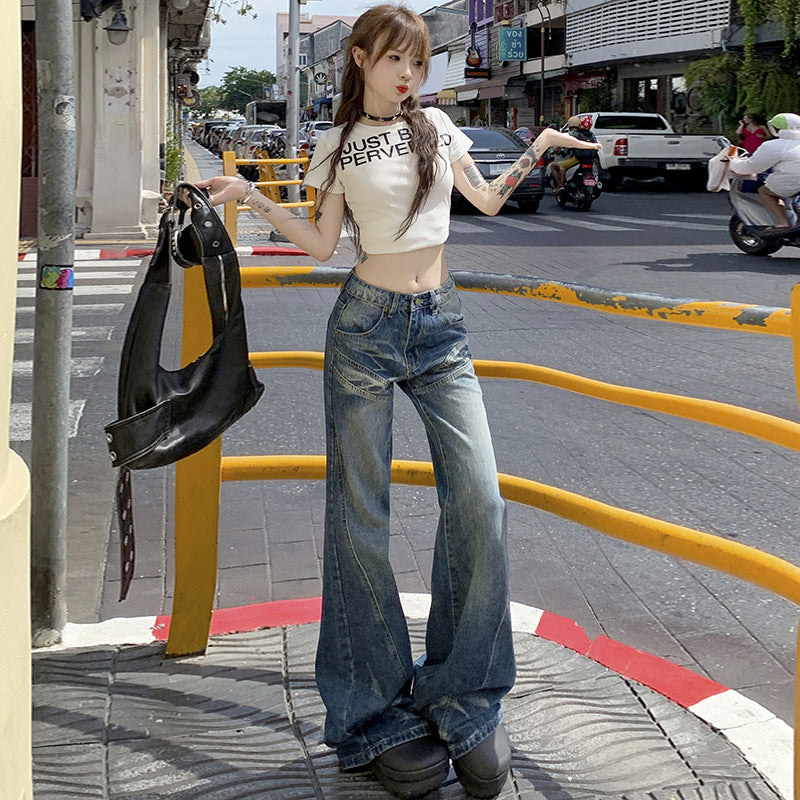 A woman standing on city sidewalk wearing white crop top with text, wide-leg faded jeans and platform shoes, holding black bag on urban street