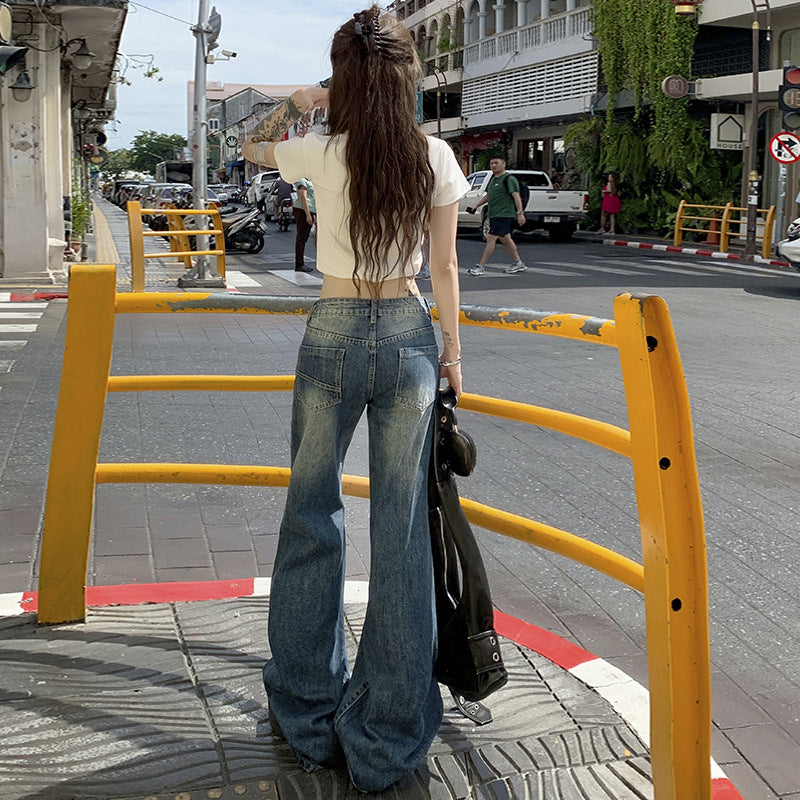 Back view of woman with long hair wearing white crop top and wide-leg faded jeans, standing at street crossing in shopping district