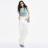 A woman with long black hair poses in a white studio. She wears a blue one-shoulder crop top with black text, white cargo pants, and blue sneakers