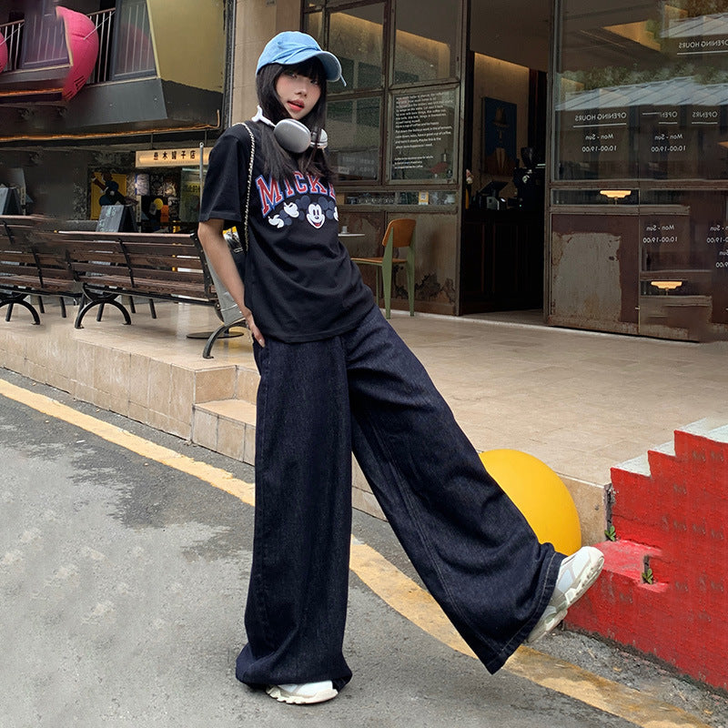 Streetwear ensemble with Mickey graphic tee, relaxed-fit dark jeans, baseball cap and white sneakers against urban backdrop