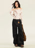 A woman is posing in a cream off-shoulder button top with black wide-leg jeans, leather choker, and brown messenger bag on light background