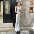 A woman wearing a white tank top with black bow details and white flare jeans poses near a storefront, carrying a small black handbag
