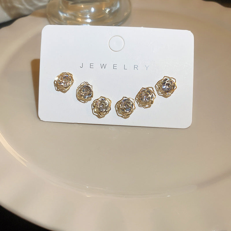  A white jewelry card displaying a set of six gold rose-shaped stud earrings with a delicate openwork design, placed on a reflective surface.