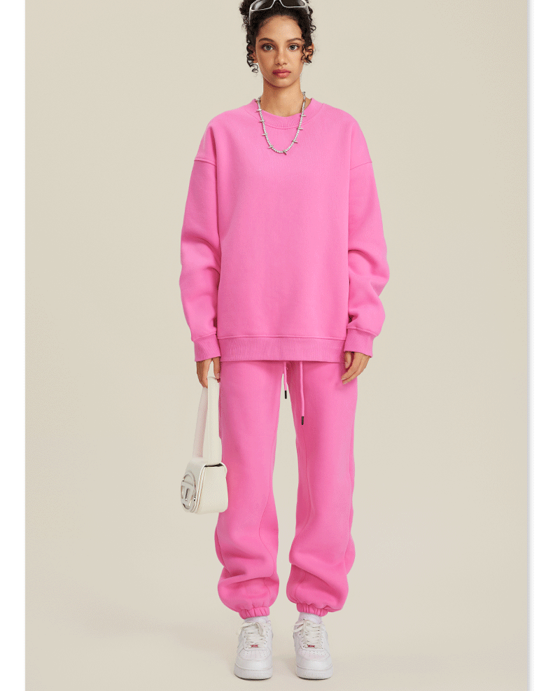 Image 1: Model wearing bright pink sweatsuit set with oversized crew neck sweatshirt and matching joggers, styled with white headband, silver necklace and white crossbody bag