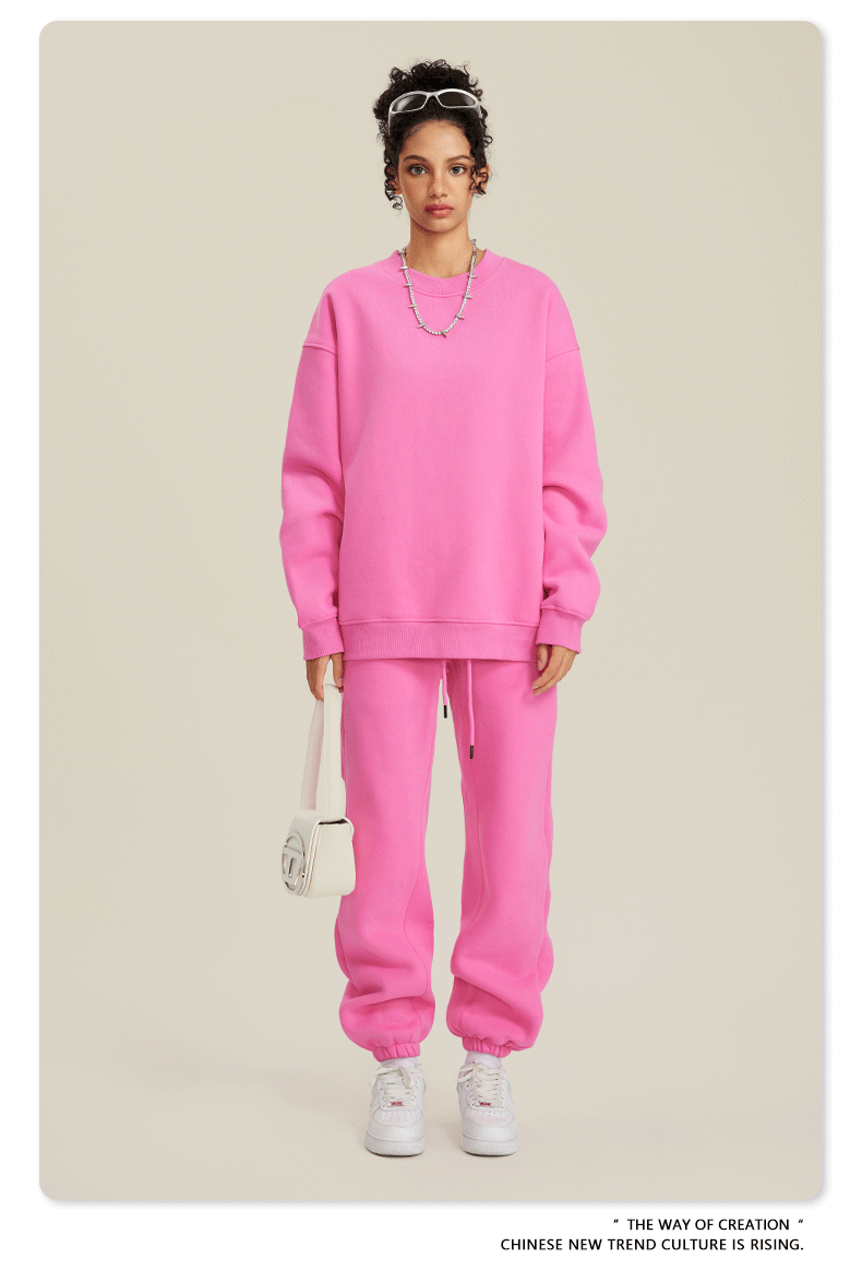 Image 1: Model wearing bright pink sweatsuit set with oversized crew neck sweatshirt and matching joggers, styled with white headband, silver necklace and white crossbody bag